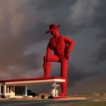 a film still of a giant red cowboy with his leg on the top of a gas stantion