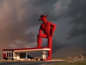 a film still of a giant red cowboy with his leg on the top of a gas stantion