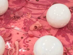 a detail of a square floor work with a white border and pink in the center. small white orbs rest on top. in the pink are smaller forms like hands, fabric, leaves, shells, and more