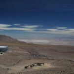 A high-altitude observatory in the Atacama Desert, Chile, surrounded by an arid landscape with distant mountains under a deep blue sky. The facility consists of a large, circular telescope structure and a few small buildings.