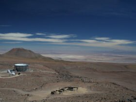 A high-altitude observatory in the Atacama Desert, Chile, surrounded by an arid landscape with distant mountains under a deep blue sky. The facility consists of a large, circular telescope structure and a few small buildings.