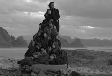 a black-and-white photograph in the Jordanian desert of a group of people in black garments perched on a stack of boxes to create a monolith-like form