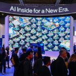 People in a blue-lit convention hall stand before a towering display with an illustration of connected communities under a sign saying "AI Inside for a New Era."