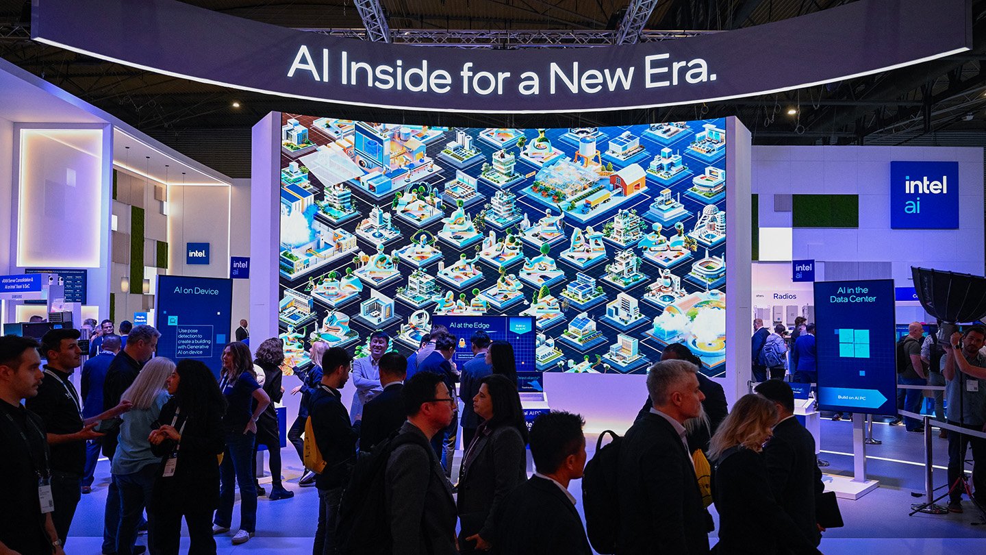 People in a blue-lit convention hall stand before a towering display with an illustration of connected communities under a sign saying "AI Inside for a New Era."