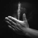 A grayscale images shows two hands clapped together, with a white jet of baby powder streaming upward from a space between the thumbs and forefingers.