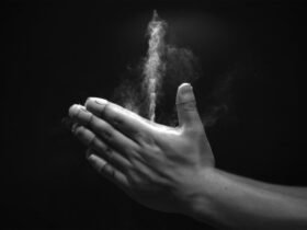 A grayscale images shows two hands clapped together, with a white jet of baby powder streaming upward from a space between the thumbs and forefingers.