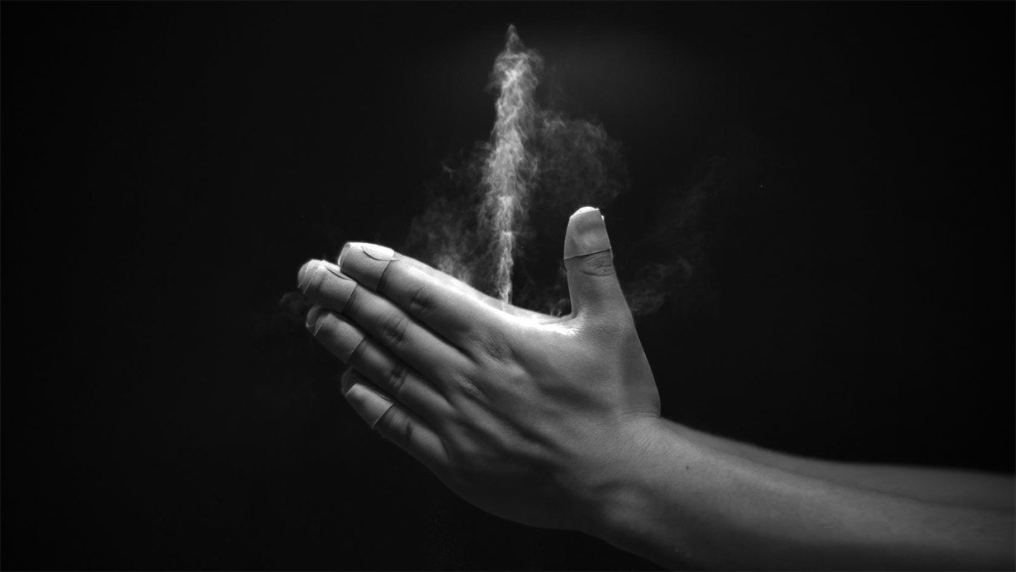 A grayscale images shows two hands clapped together, with a white jet of baby powder streaming upward from a space between the thumbs and forefingers.