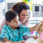 A girl, wearing patient attire, on a hospital bed pets a therapy dog. The girls