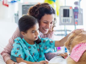 A girl, wearing patient attire, on a hospital bed pets a therapy dog. The girls