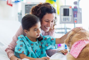 A girl, wearing patient attire, on a hospital bed pets a therapy dog. The girls