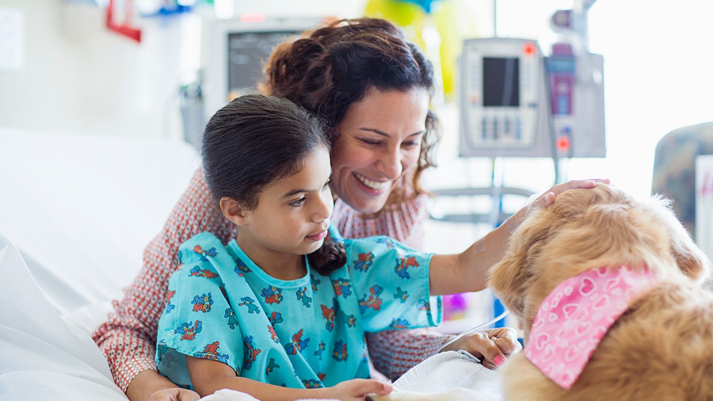 A girl, wearing patient attire, on a hospital bed pets a therapy dog. The girls