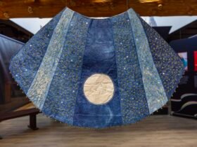 a barkcloth tapestry installation with indigo-dyed panels in different shades, sewn together to create a ray-like effect, with a metallic circle in the bottom center