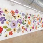 a large, full-wall installation of numerous flower painting cutouts that resemble giant pressed flowers all along a white wall