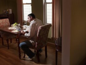 A man sitting alone at a dining table in a dimly lit room, reading something, with a glass of red wine and a partially eaten meal in front of him.