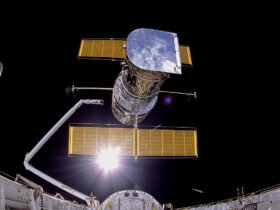 The Hubble Space Telescope being deployed from a space shuttle into orbit, with solar panels extended against the black backdrop of space and the sun shining brightly behind the observatory.