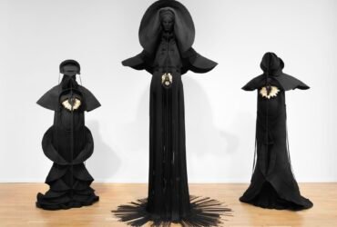 Installation view of three sculptures made from black cord, thread, and metal in the form of figures or long, dramatic garments