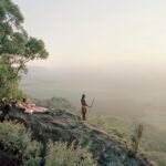 a Masai warrior stands on a hillside, looking out over a hazy landscape, with a pink picnic blanket on the rocks behind him
