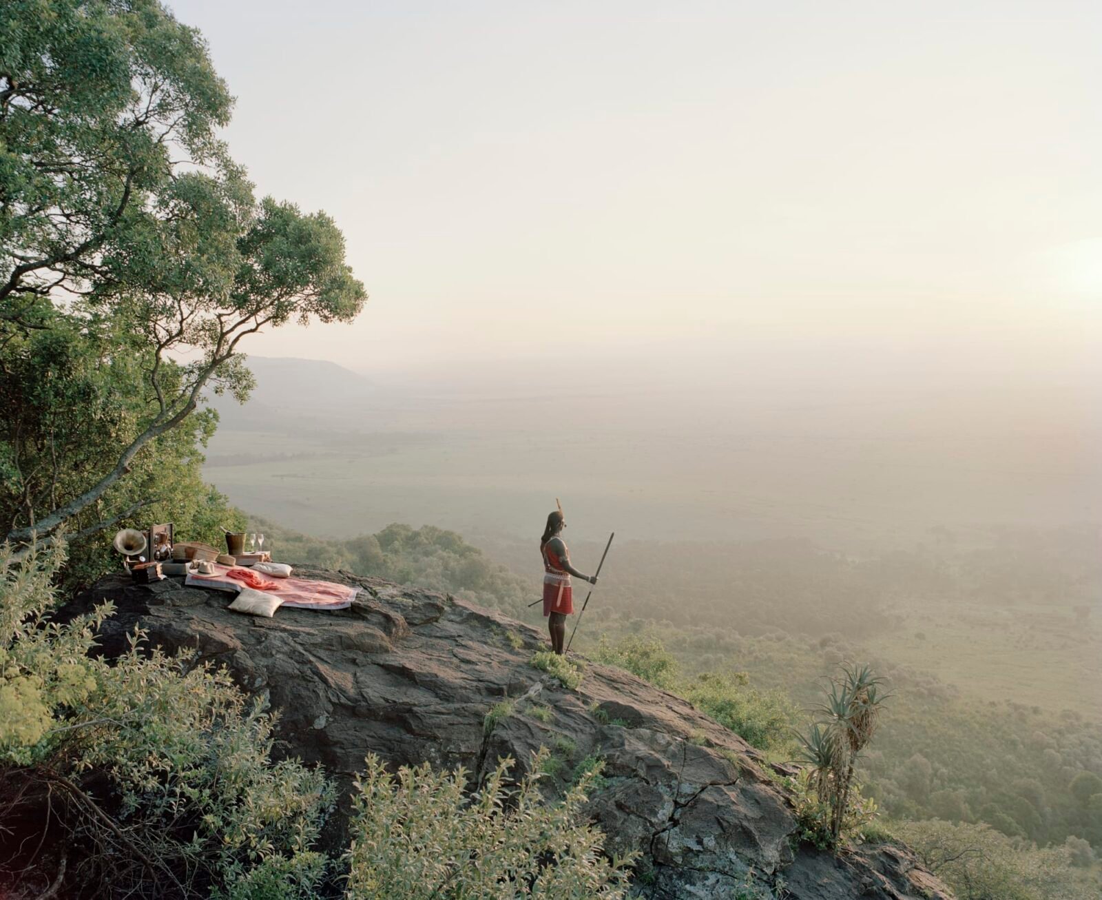 a Masai warrior stands on a hillside, looking out over a hazy landscape, with a pink picnic blanket on the rocks behind him