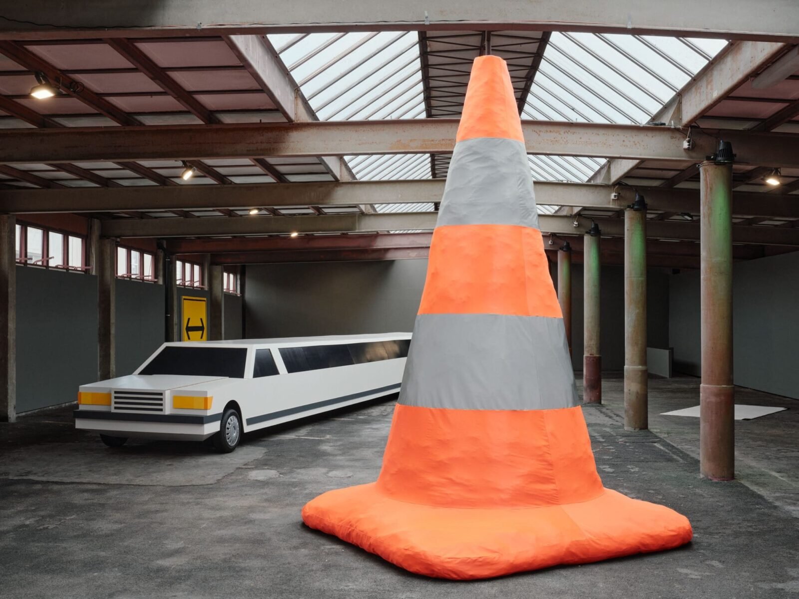 a cavernous gallery space with a large sculpture of a very long stretch limo made of cardboard and a huge orange safety cone in the foreground