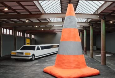 a cavernous gallery space with a large sculpture of a very long stretch limo made of cardboard and a huge orange safety cone in the foreground