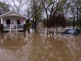 Muddy-looking water surrounds a car and house, creeping up toward the car windows and the steps of the front porch.