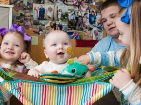 A smiling baby who got a personalized gene therapy is sitting in a multi-colored striped bouncy seat is surrounded by three siblings. A girl with long red hair and purple bows smiles at the camera on the left. A boy in a blue shirt watches as a girl with long blonde hair held back by a blue bow offers the baby a stuffed toy.