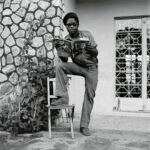 a black-and-white photograph in a square format of a Black man standing outside of a house, wearing fashionable 1970s apparel, standing with one leg up on the seat of a chair and holding a magazine