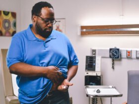 A man injects himself in the stomach with a "pen" containing a GLP-1 drug. The man has dark skin and hair, is wearing glasses and wearing a blue polo-style shirt. He is lifting his shirt to expose the spot on his stomach where he will inject the medication. He appears to be in a doctor