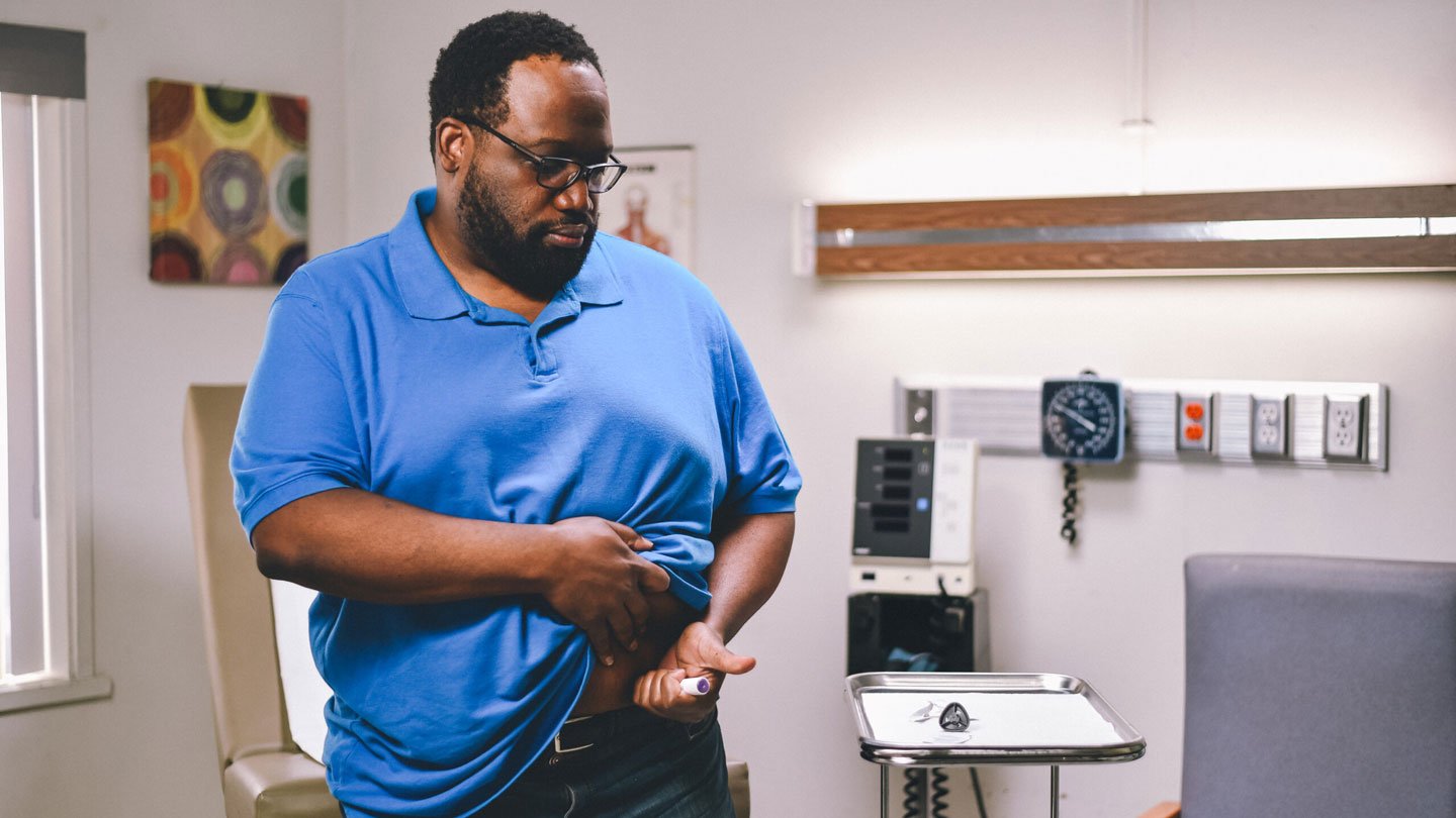 A man injects himself in the stomach with a "pen" containing a GLP-1 drug. The man has dark skin and hair, is wearing glasses and wearing a blue polo-style shirt. He is lifting his shirt to expose the spot on his stomach where he will inject the medication. He appears to be in a doctor