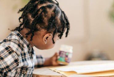 A young toddler wearing a flannel shirt sits at a table and holds a jar of food.