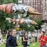 a procession of people in a park carry three puppets of sturgeons on poles