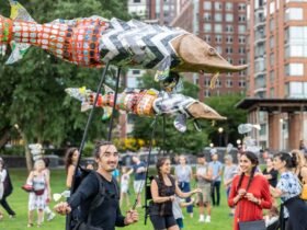 a procession of people in a park carry three puppets of sturgeons on poles