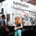 a white woman stands in front of a graffitied sanitation truck talking to a black man
