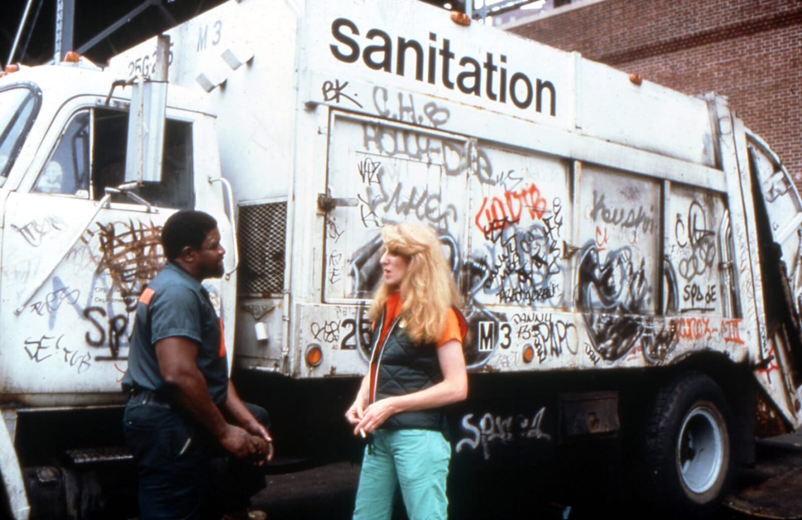 a white woman stands in front of a graffitied sanitation truck talking to a black man