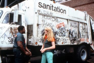 a white woman stands in front of a graffitied sanitation truck talking to a black man