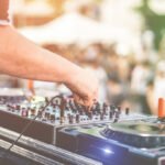 A close-up of hands at a DJ table with a blurred summertime crowd in the background