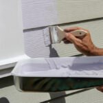 A close-up from the elbows down of a person painting the siding of a house. One hand holds a paintbrush while the other holds a tray of white paint