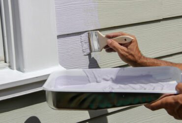 A close-up from the elbows down of a person painting the siding of a house. One hand holds a paintbrush while the other holds a tray of white paint