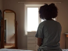 A woman with her back to the camera sits on a bed in a dimly lit room looking out the window